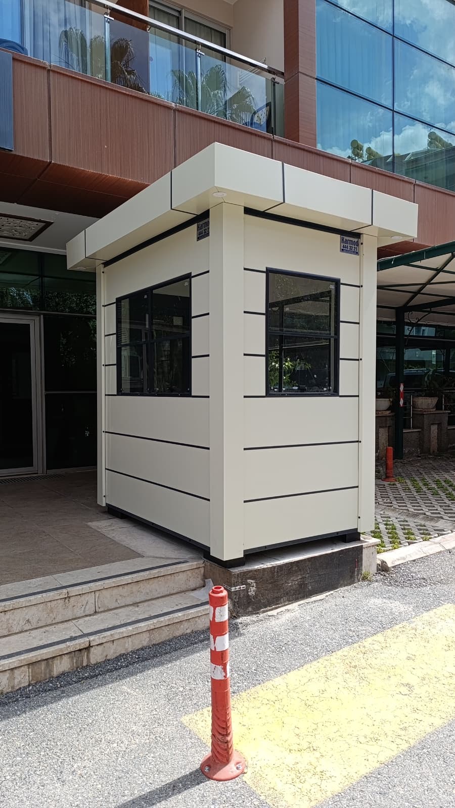 Cream composite-clad gatehouse cabin with a deep overhanging roof, standing at the entrance of a modern office building.