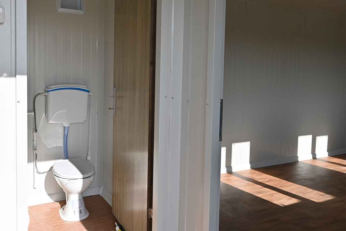 WC compartment inside a standard container unit, with a close-coupled toilet and a partition door to the main room.