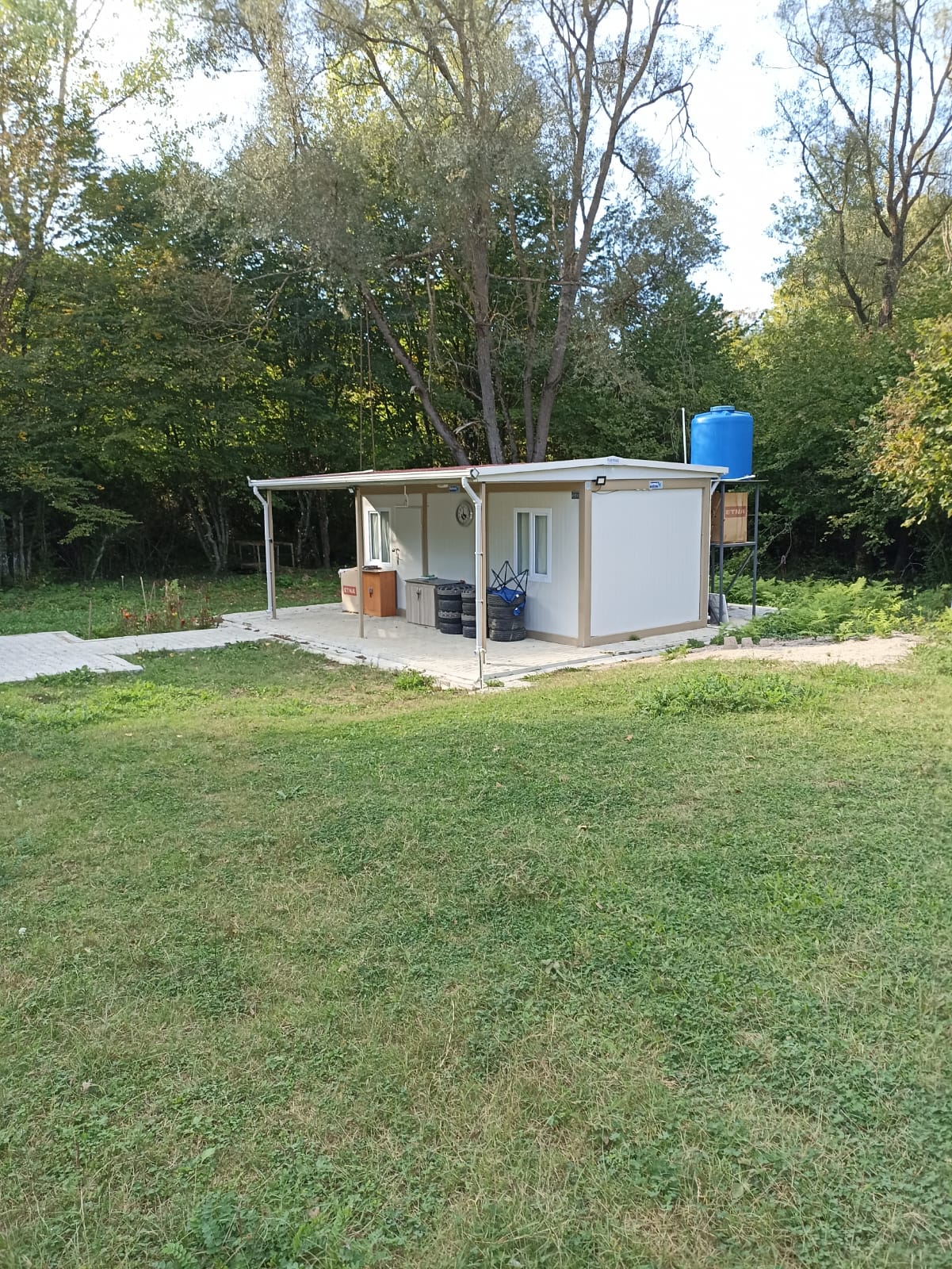 Small white container house with a canopied porch and a raised water tank, in a mown clearing against dense woodland at Sevindikli.
