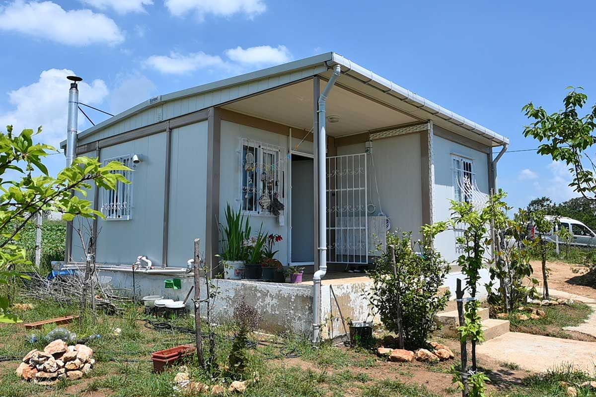 Container home with a canopied porch, a flue and grilled windows on a rural smallholding, with young fruit trees around it.
