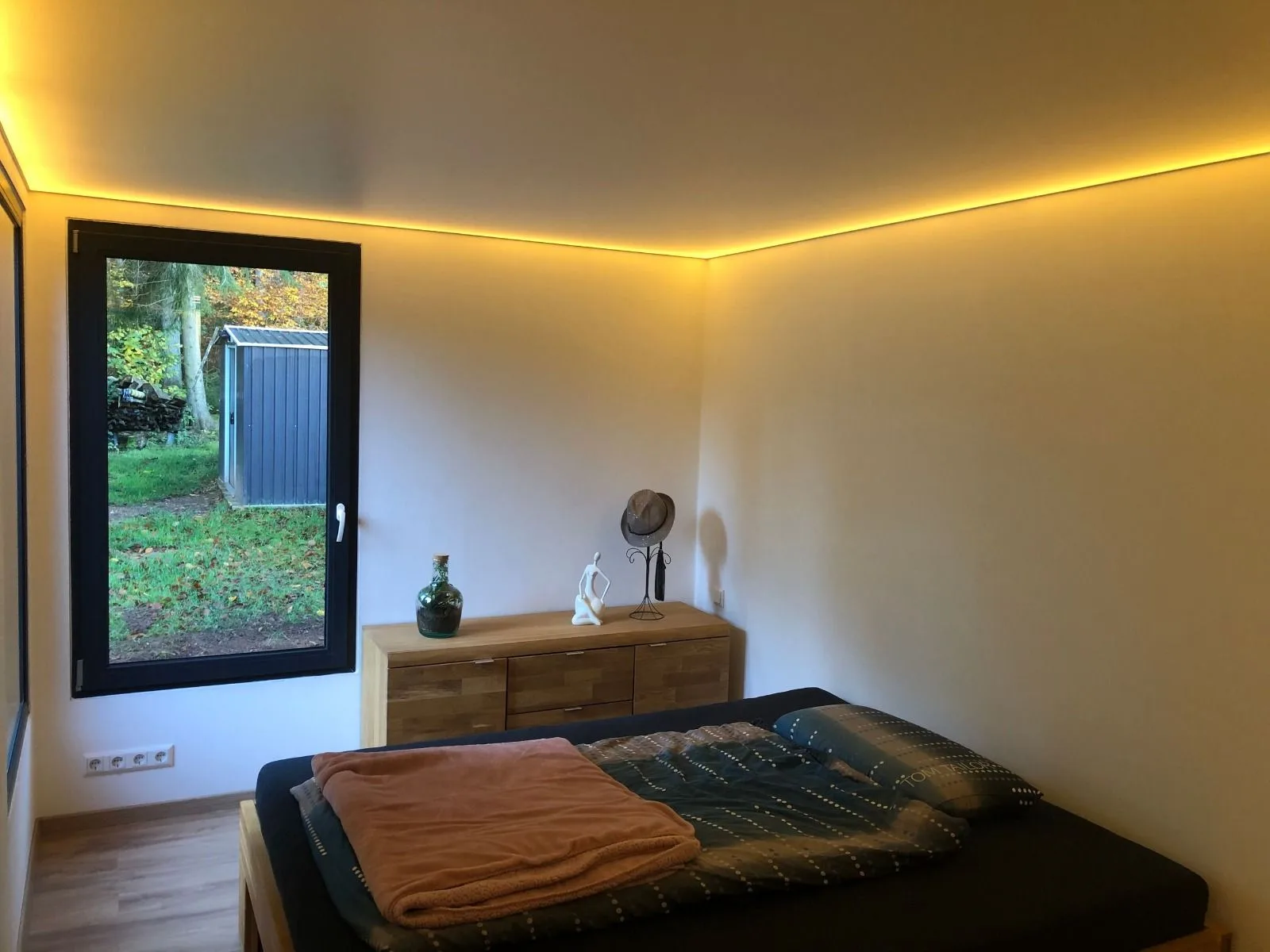 Container house bedroom with concealed cove lighting, a double bed and an oak sideboard, the window looking onto autumn woodland.