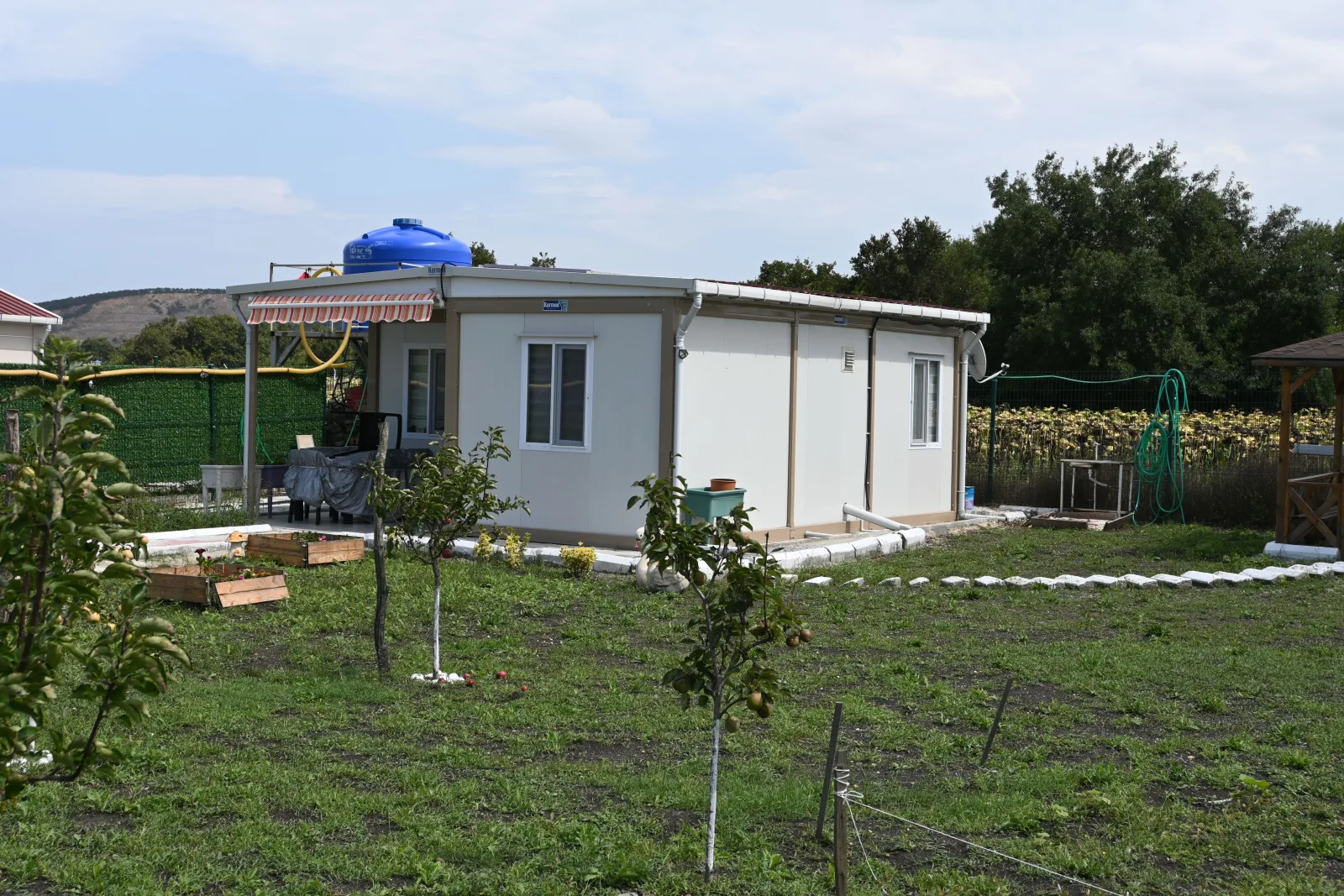 White container house with a striped awning and a rooftop water tank, set in a cultivated garden at Akalan Catalca.