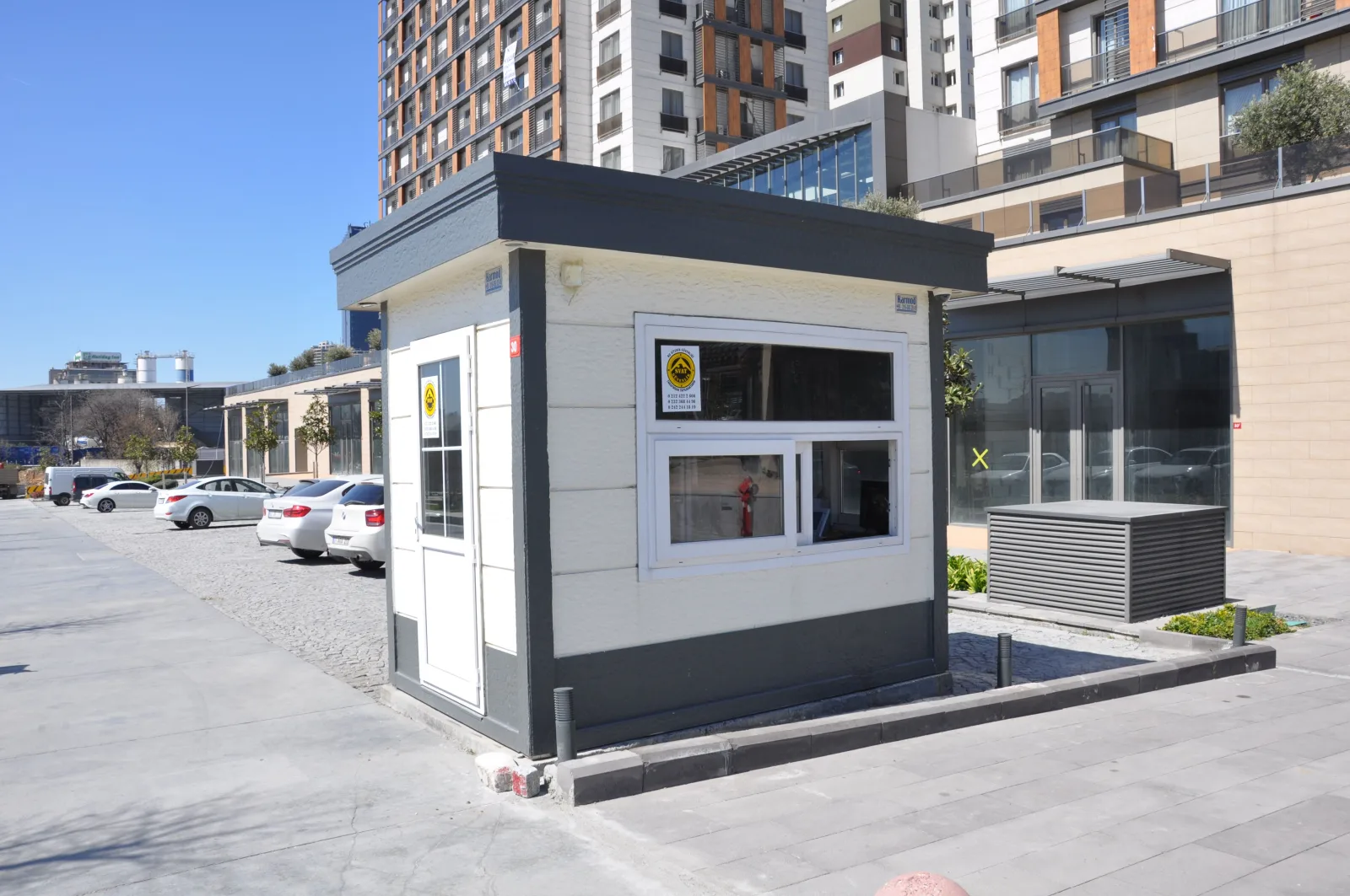 White and anthracite Metro City security cabin on the paved forecourt of the Double Park development, with apartment towers behind.