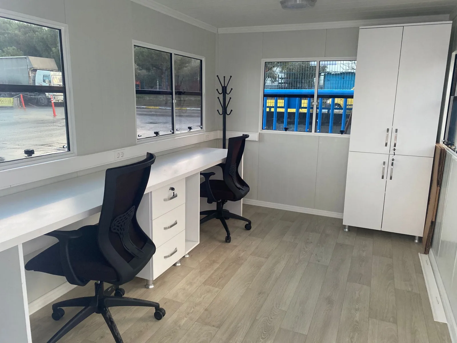Interior of a Metro City site office with two desks along a continuous worktop, office chairs, a tall cupboard and wood-effect flooring.
