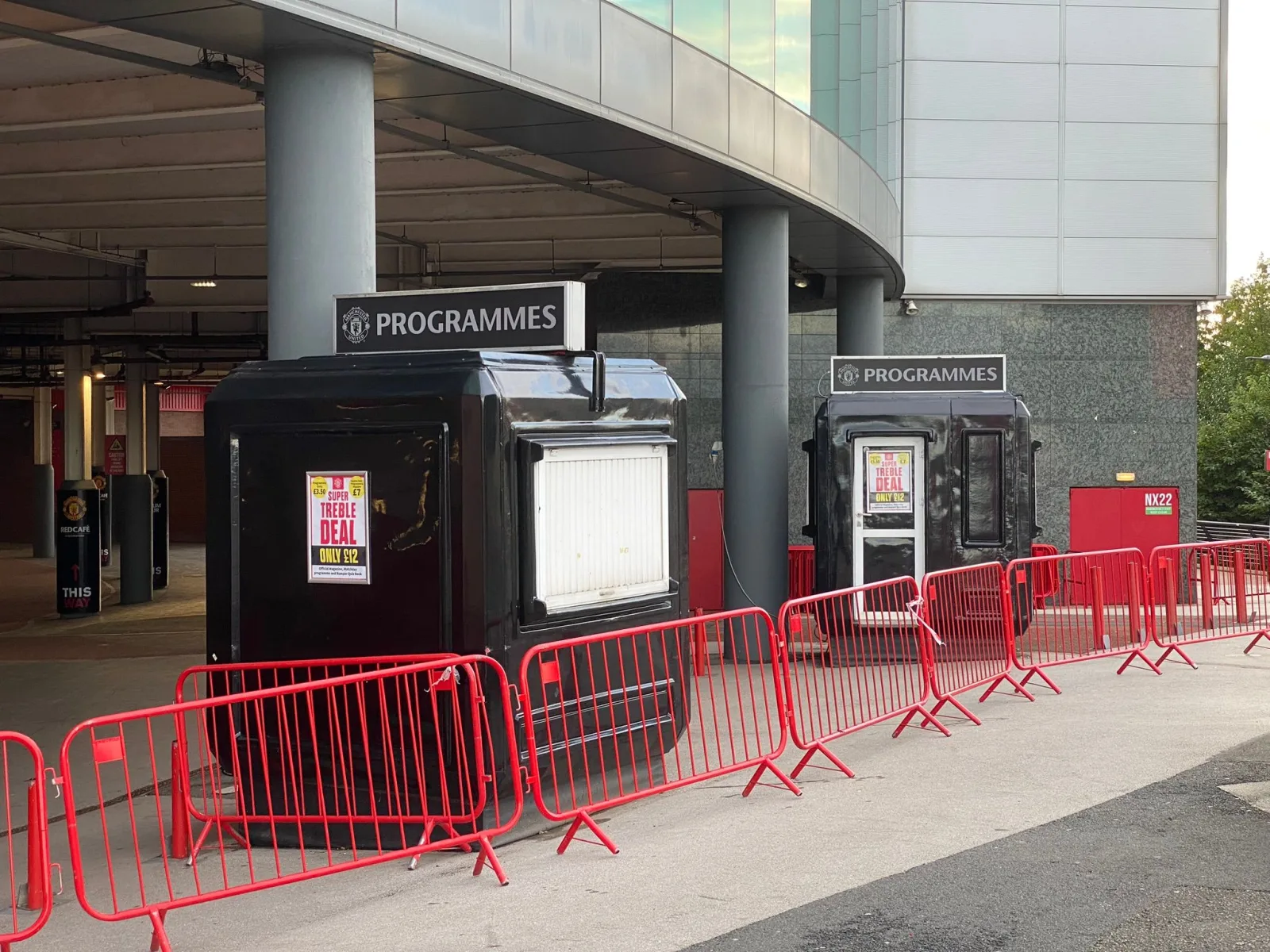 Two black Manchester United programme kiosks under the stadium concourse canopy at Old Trafford, behind red crowd barriers.
