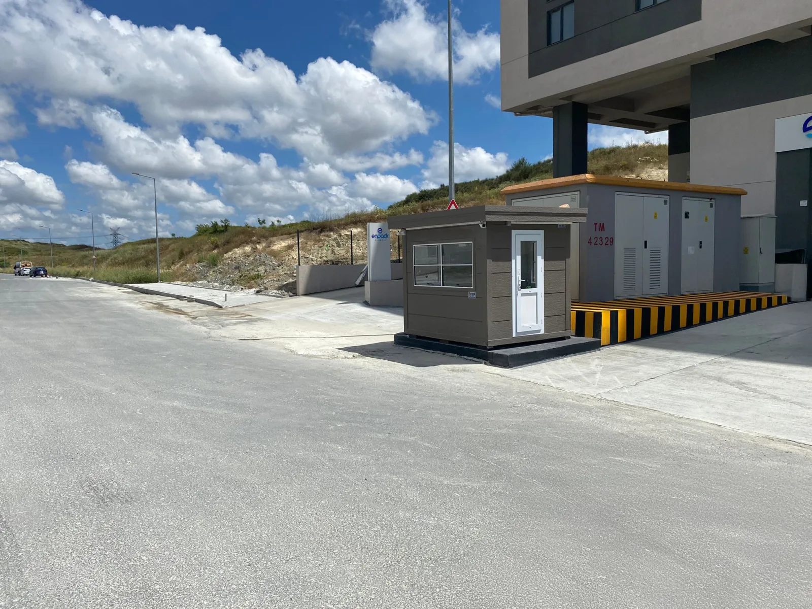 Taupe Metro City cabin with a white door beside a substation enclosure at the entrance to a factory site.