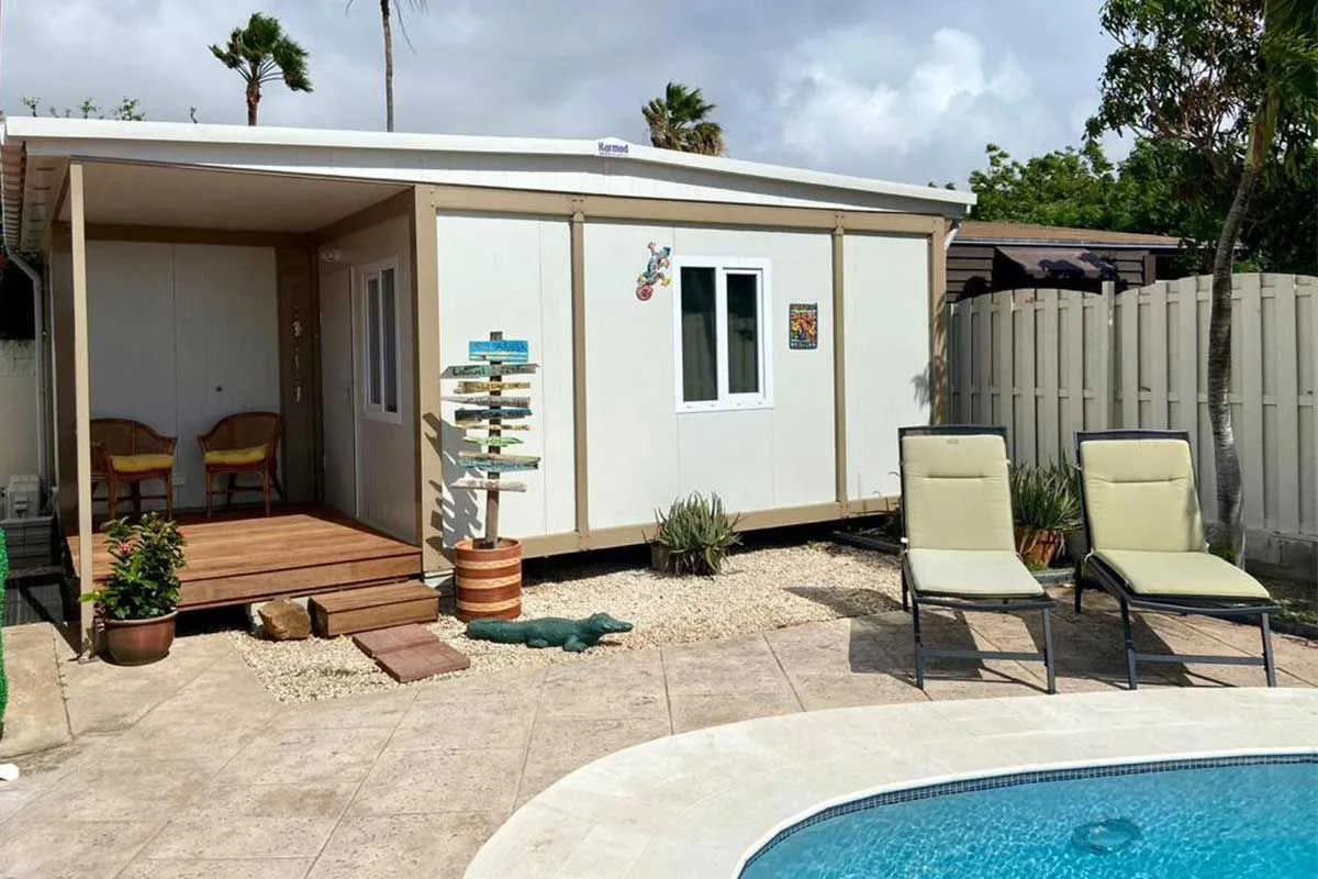 Container guest suite with a timber deck and a covered seating area beside a swimming pool, with palm trees behind.