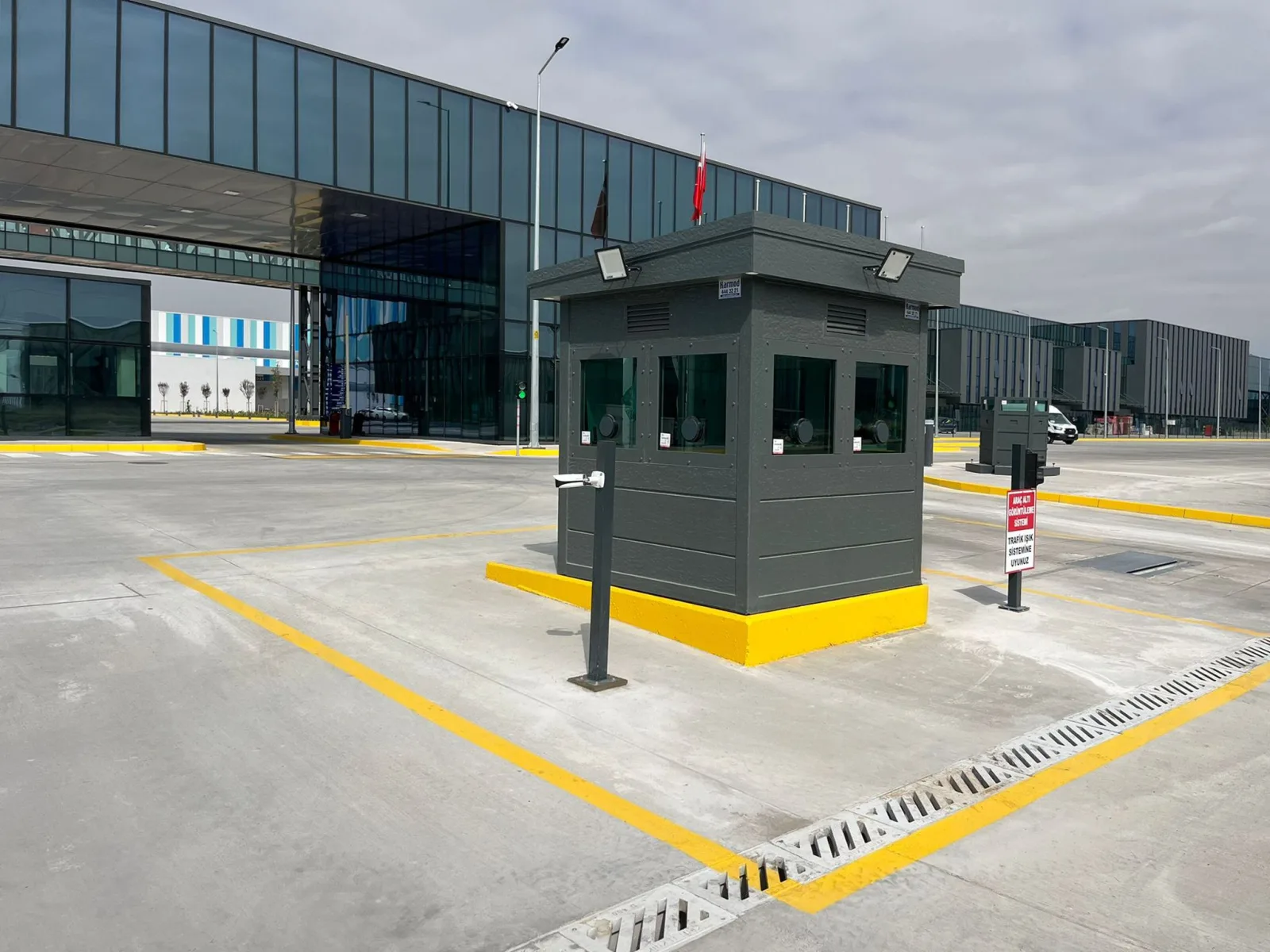 Dark grey armoured security cabin on a yellow-painted plinth, controlling the vehicle gate of a large manufacturing plant.