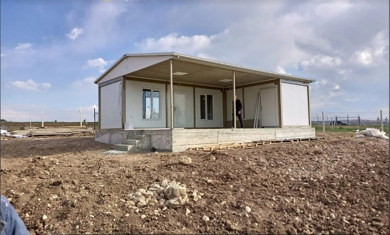 Container house with a canopied terrace being installed on a levelled plot, with timber stacked alongside.