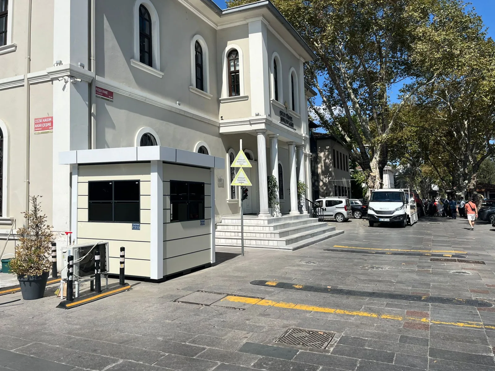 Cream and white composite cabin with a wide roof canopy, set on paving beside a historic foundation building on an Istanbul street.