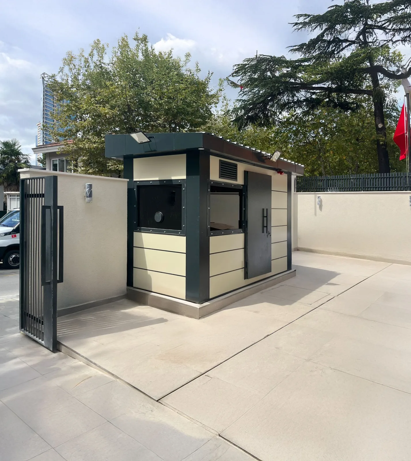 Cream and dark green armoured composite cabin with a ballistic window and armoured door, on a paved forecourt beside a gate.