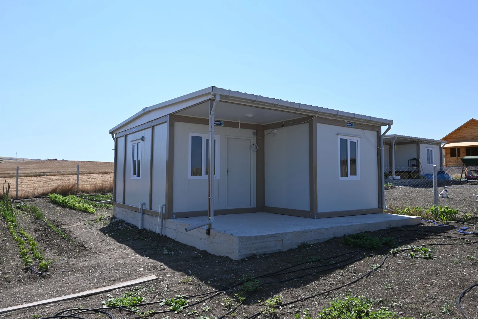 Container house with a canopied terrace at the edge of harvested farmland near Ankara, with irrigation lines in the foreground.