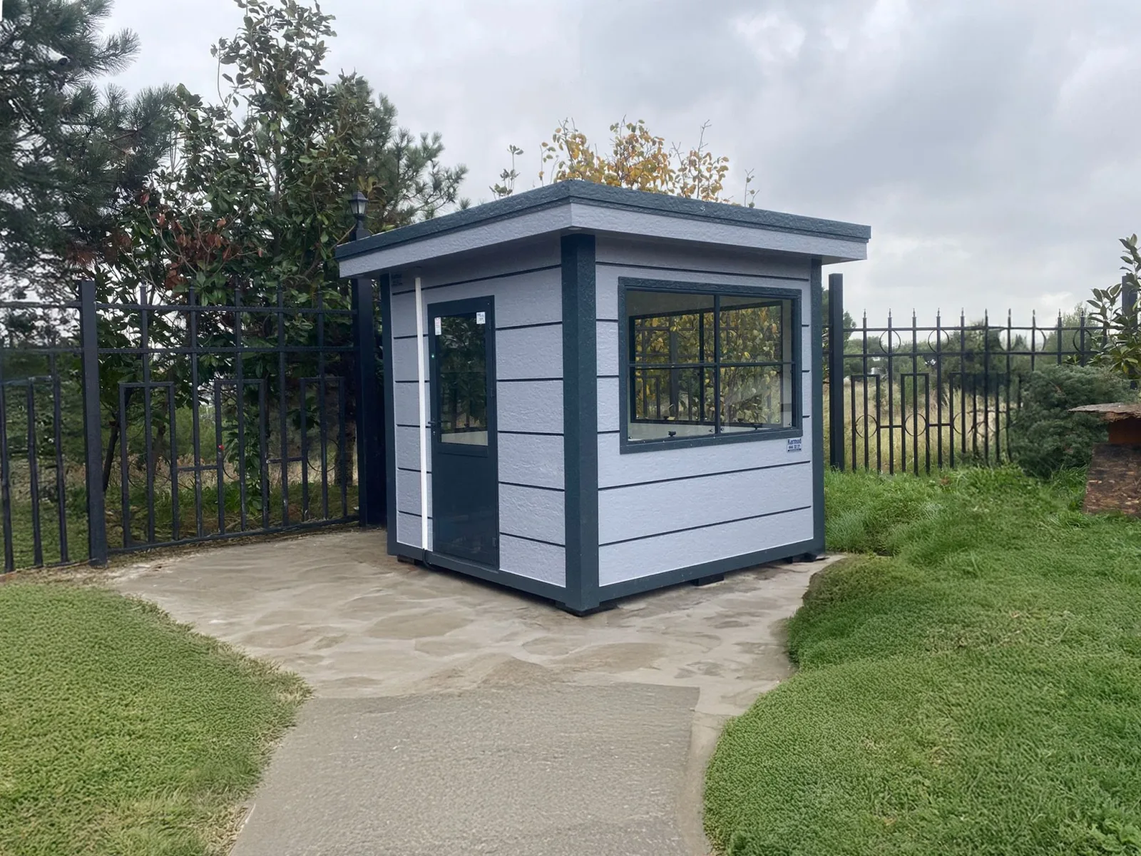 Pale grey Metro City cabin with dark trim, standing on a paved path beside railings and planting on a university campus.