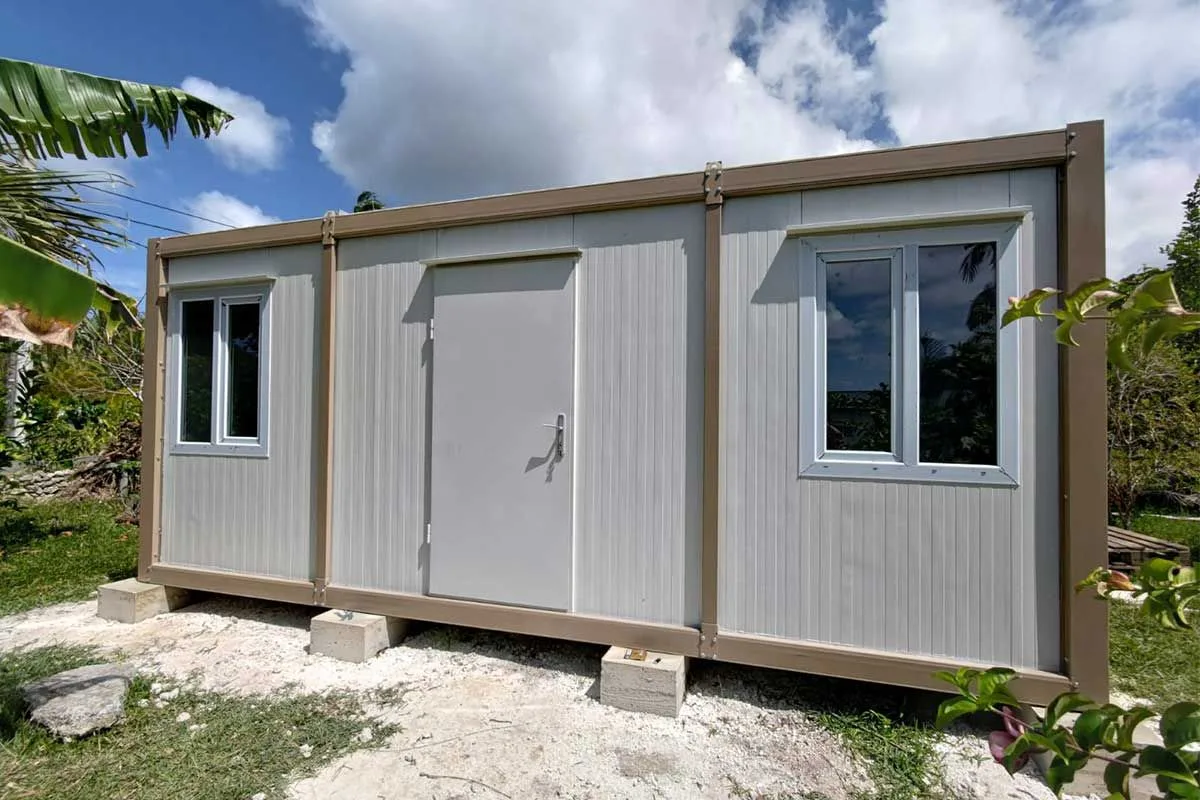 Grey and beige container unit raised on concrete footings in a tropical garden, surrounded by banana and palm foliage.