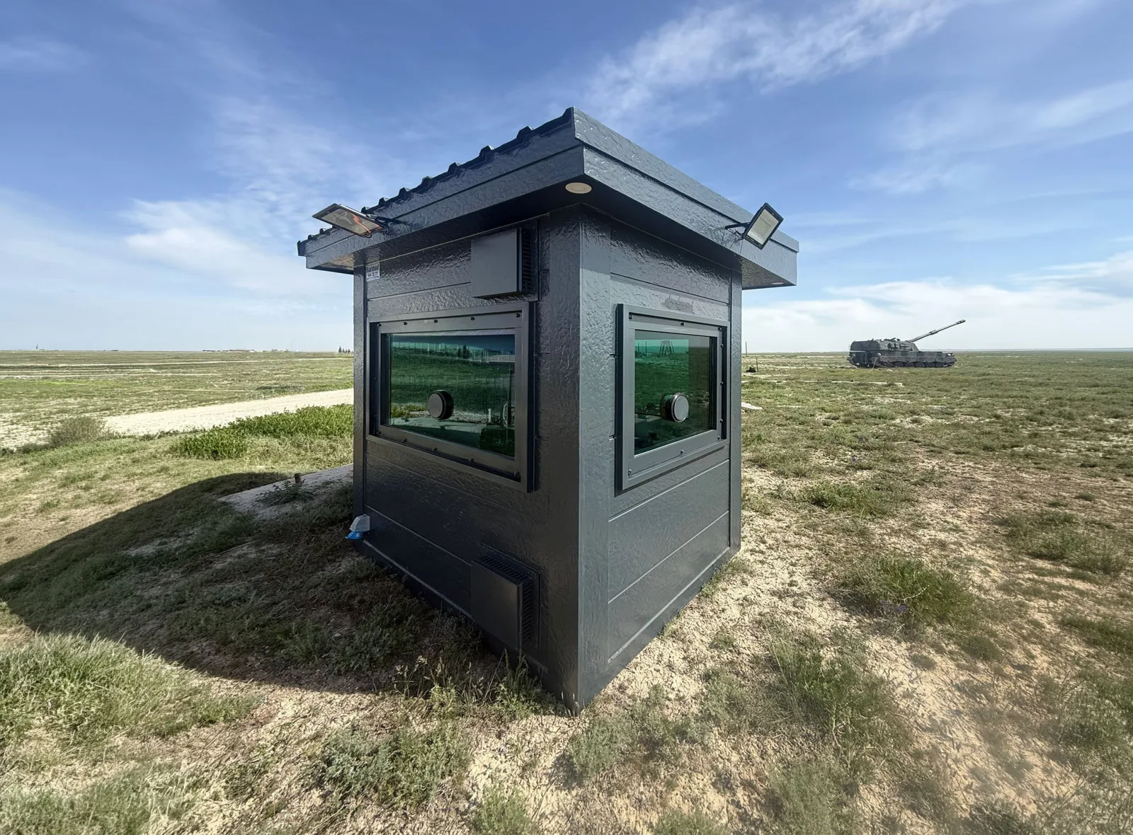 Dark textured armoured cabin with ballistic glazing and firing ports, standing alone on open grassland.