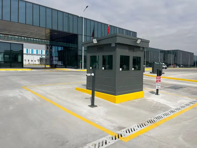 Grey armoured gatehouse on a raised plinth controlling the vehicle entrance to a manufacturing plant