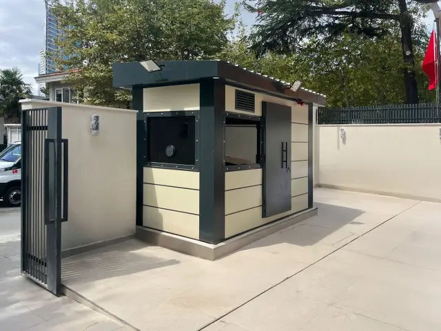 Cream and dark grey composite-clad armoured cabin with a steel door on a paved private forecourt beside a gate